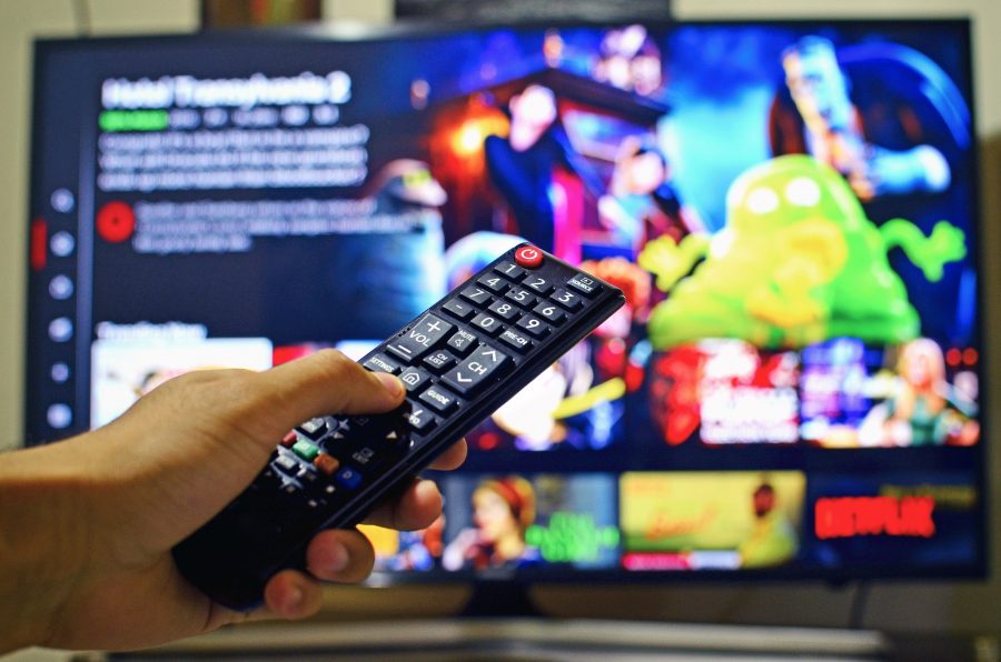 A hand holding a remote control with Netflix on a TV in the background